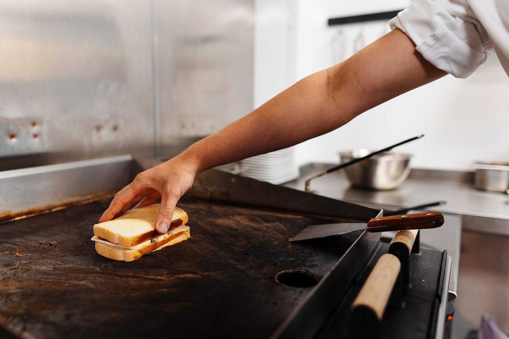 A professional food photographer capturing a close-up of a chef assembling a sandwich on a commercial griddle in a busy kitchen.