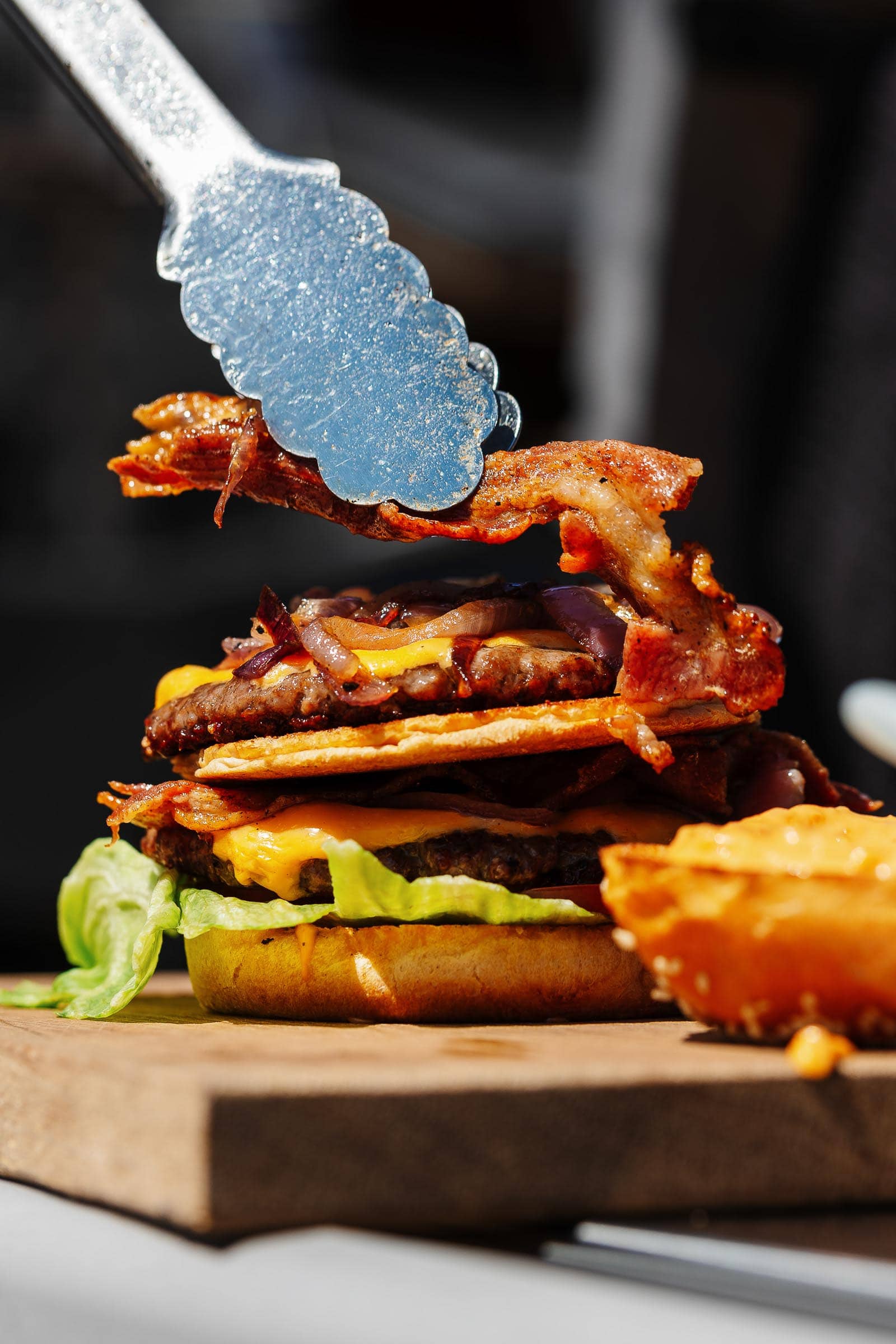 A slice of bacon being placed onto a large burger outdoors