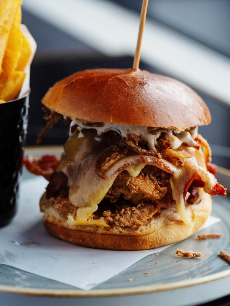 Juicy gourmet burger with crispy bacon, melted cheese, shredded onions, and a brioche bun, served with golden fries. Perfect for food photography and showcasing delicious UK comfort food.