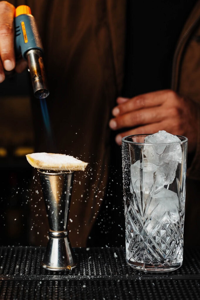 Flame torch caramelising a dessert on a metal stand with sugar, next to a glass of ice cubes, in a professional food photography setting.