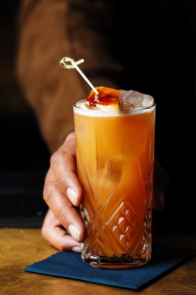Vibrant orange cocktail in a tall, cut-glass garnished with a cherry and ice, held by a bartender with a brown jacket, on a wooden bar counter with a blue napkin, showcasing professional food photography for Rutland food photography.