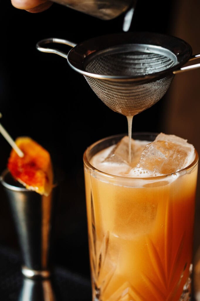 Vibrant orange cocktail with ice in a tall glass, being strained through a fine mesh sieve, on a dark background.