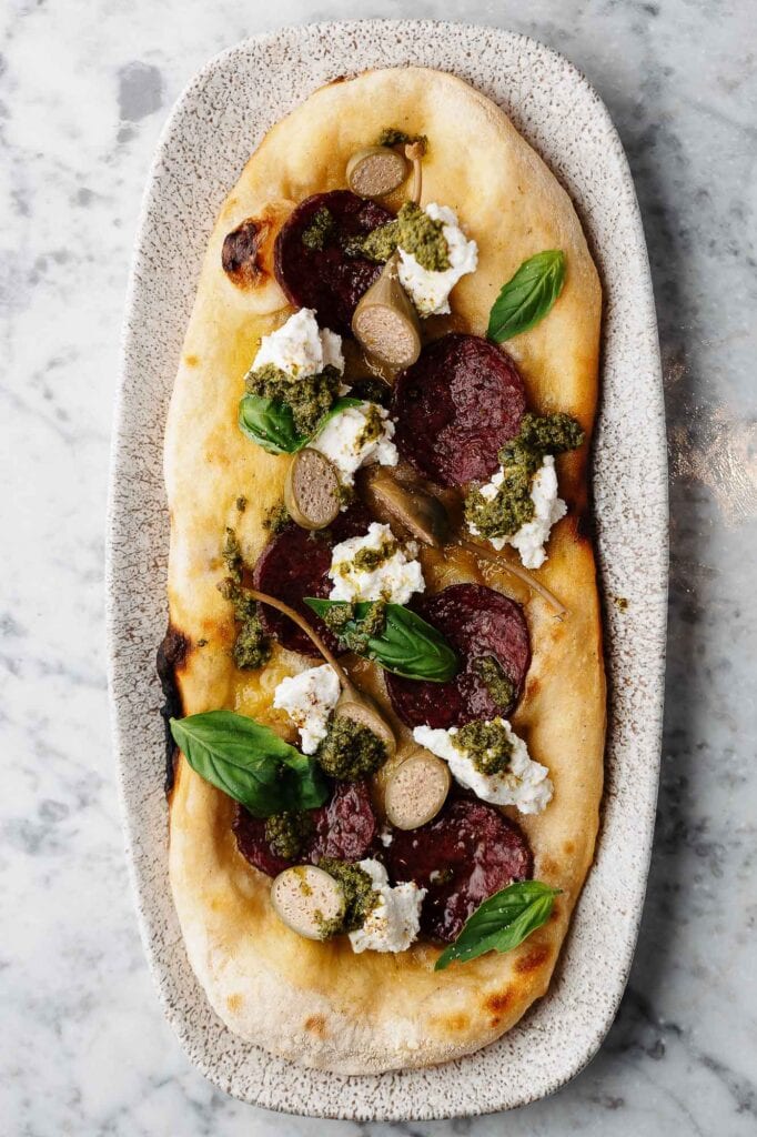 Fresh gourmet pizza with cured meats, ricotta cheese, basil, and pesto on a rustic plate, perfect for food photography showcasing delicious restaurant dishes in Rutland.