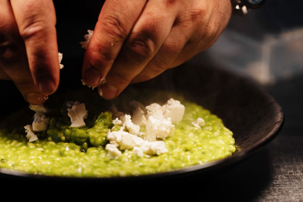 Creamy green risotto with crumbled cheese being garnished by a chef's hand, Food photographer in Rutland, vibrant food styling, professional kitchen setting, highlighting fresh ingredients and detailed textures.