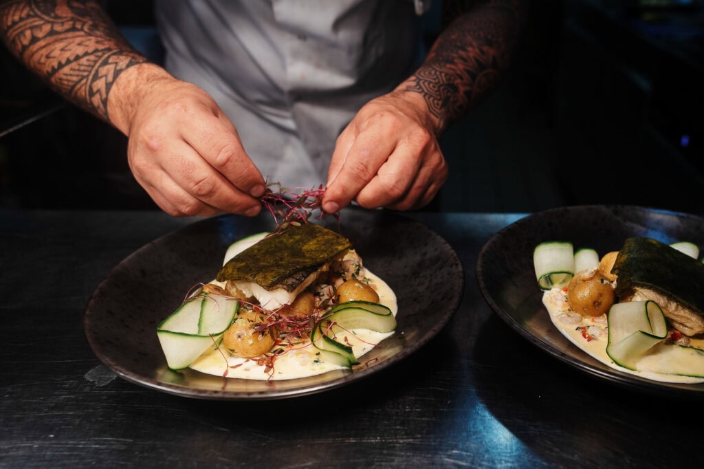 Elegant seafood dish being expertly garnished with microgreens by a tattooed chef, showcasing professional food photography for Rutland Food Photographer on a dark table.