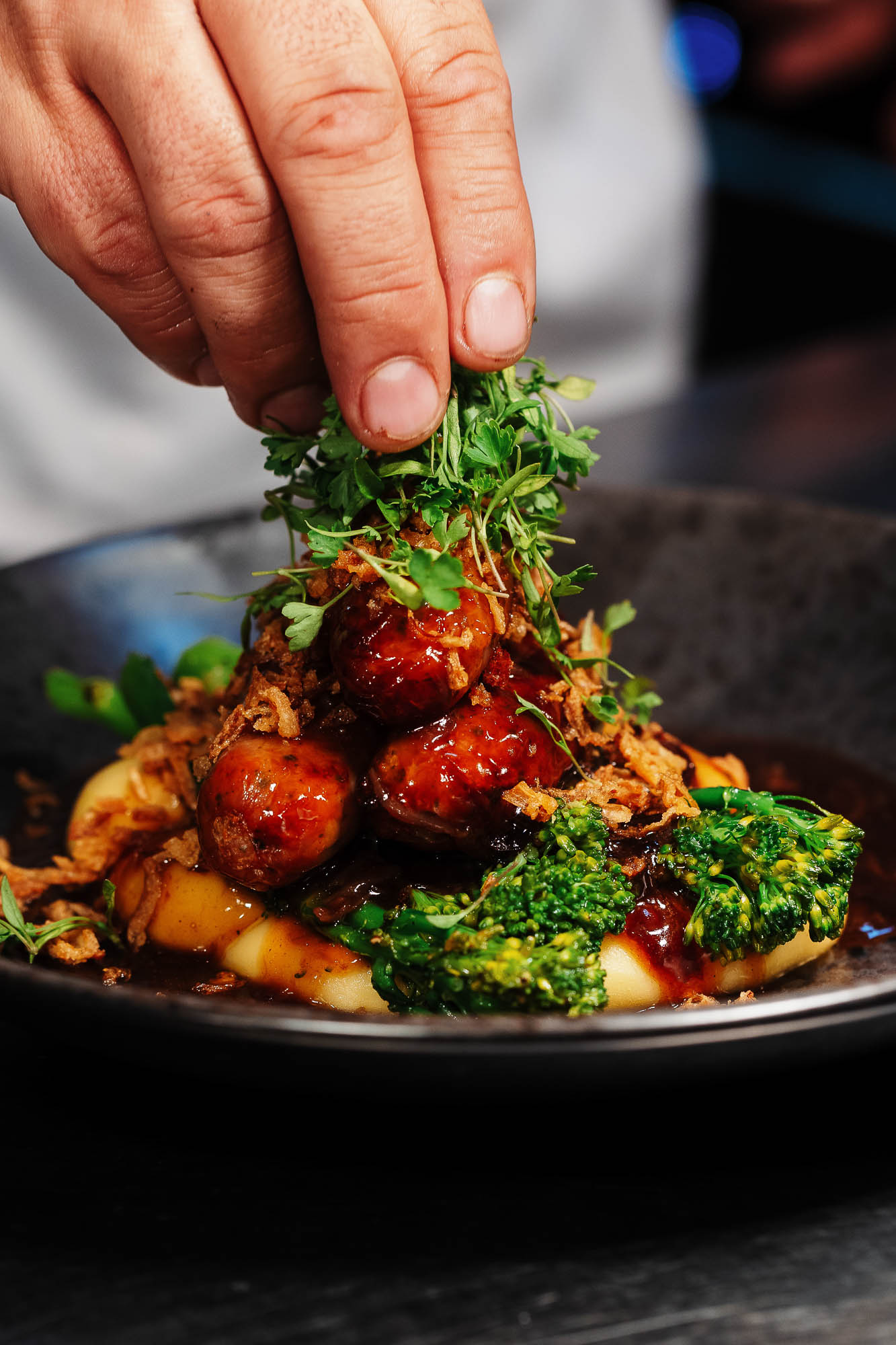 Hotel Food photographer Succulent sausages topping creamy mashed potatoes with fresh broccoli and herbs, chef garnishing a gourmet dish, professional food photography, Rutland Food Photographer, clean plate catering, vibrant food styling, fine dining presentation.