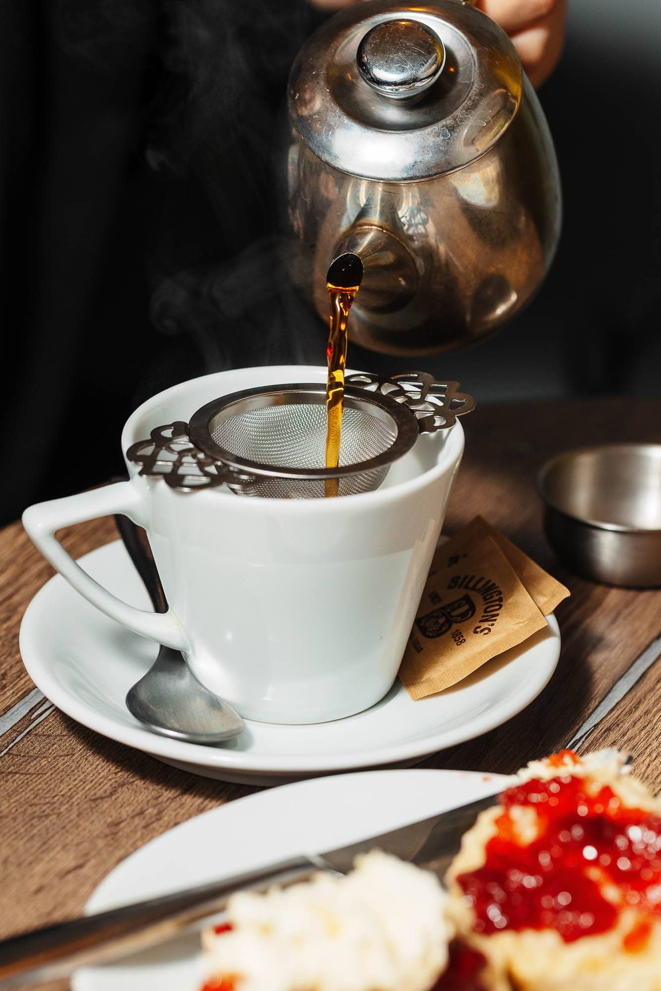 Rich coffee being poured through a metal strainer into a white mug, with a wooden table and dessert in the foreground; professional Rutland food photographer showcasing coffee and dessert pairing.