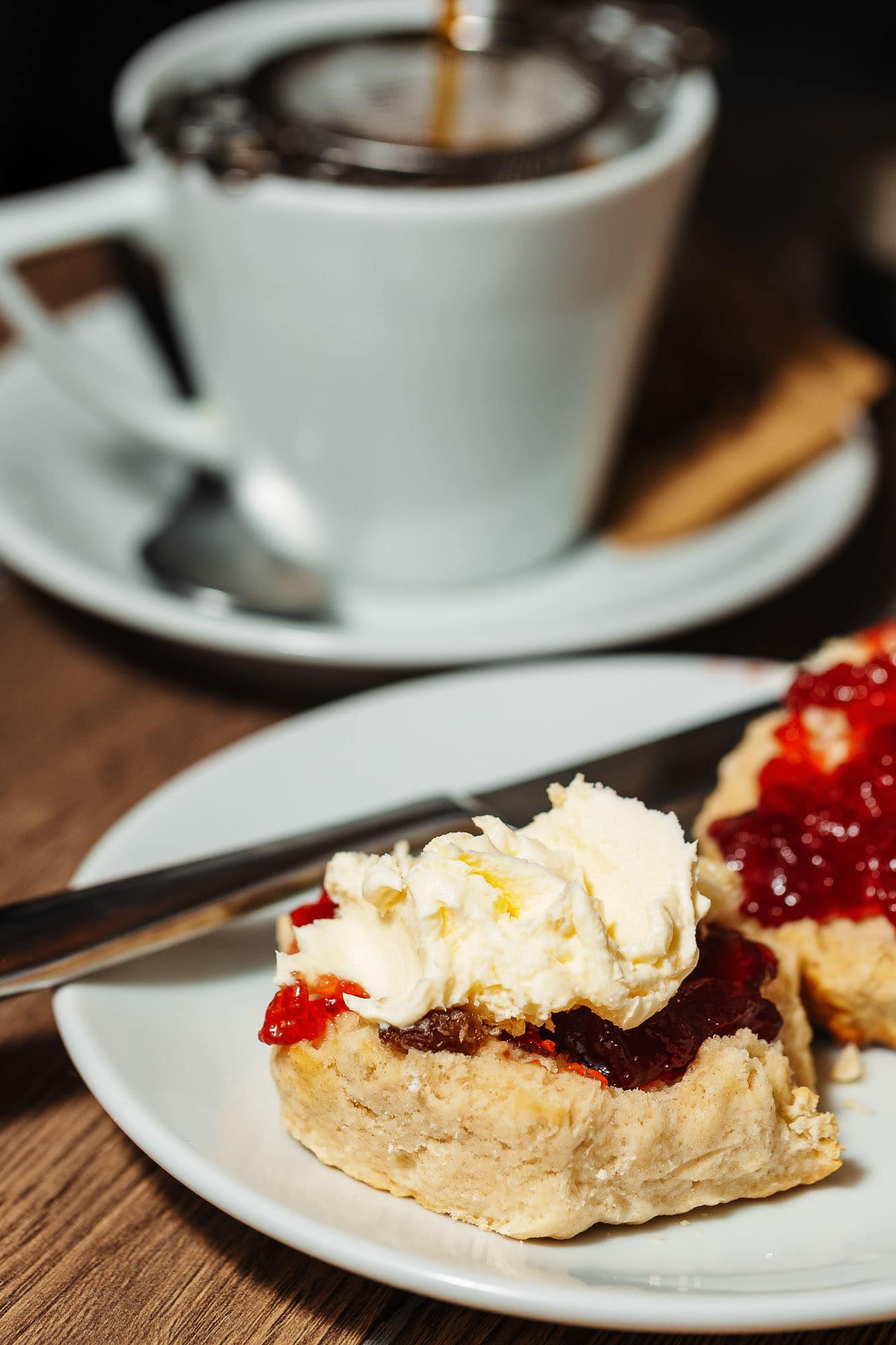 Delicious homemade cherry and cream scone on white plate with fresh cream and cherry jam, served with hot beverage in a white mug, in a cosy Rutland food photography setting.