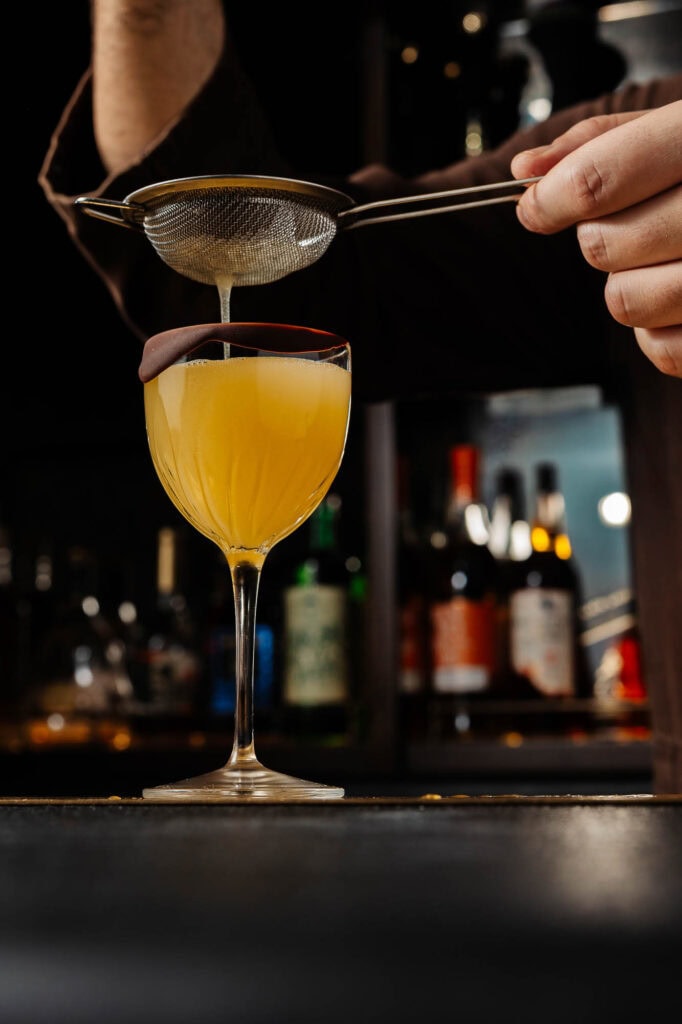 1. Elegant yellow cocktail being prepared with fine strainer, bar setting, professional bartending, drinks photography, Rutland food photographer, stylish beverage presentation, beverage styling.