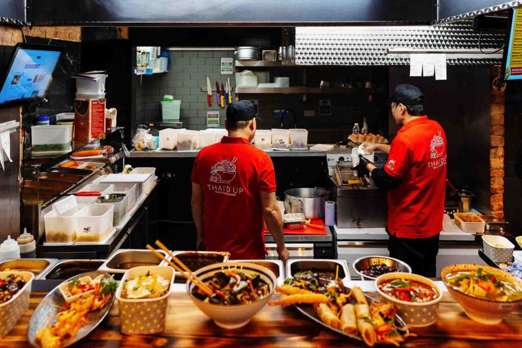 Bright Thai street food kitchen with chefs preparing dishes in a busy restaurant setting.