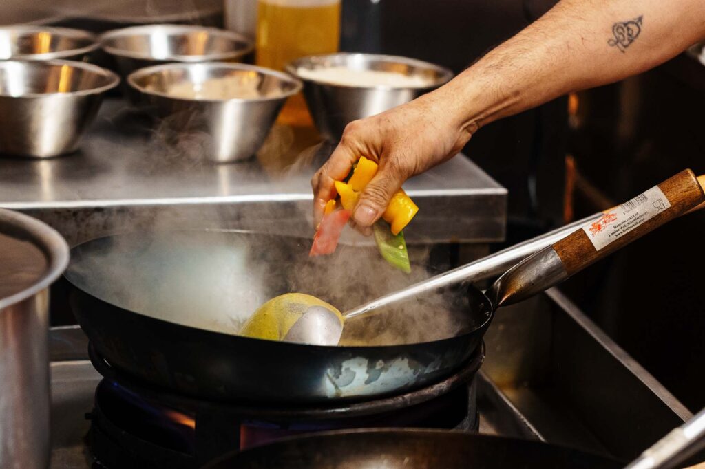 Professional food photographer capturing a chef's hand adding ingredients to a hot pan.