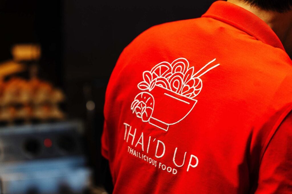 Close-up of a person wearing a red uniform with a Thai food logo featuring a bowl of noodles and chopsticks, representing a professional Thai food photographer in Rutland.