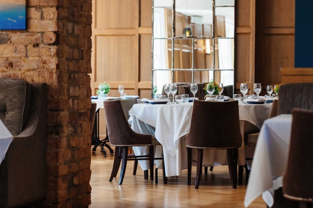Elegant restaurant interior with round dining tables set with wine glasses, cutlery, and floral centrepieces, featuring exposed brick walls and modern decor, ideal for fine dining experiences.