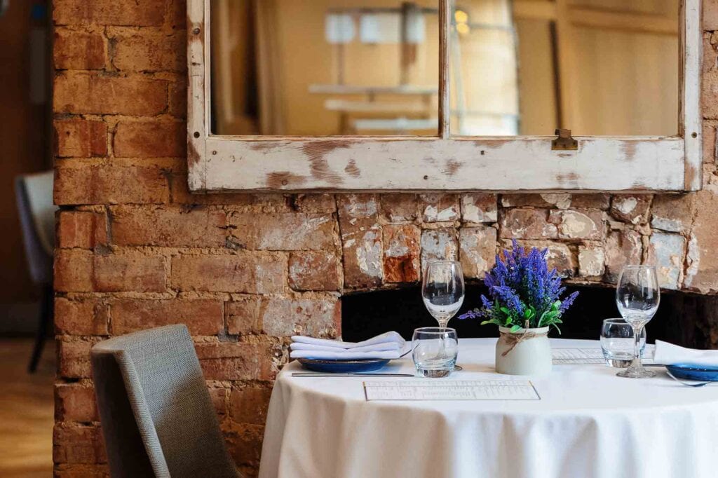 A stylish dining table set with wine glasses, white napkins, and a vibrant purple flower arrangement, situated against an exposed brick wall in a cosy, rustic restaurant environment.
