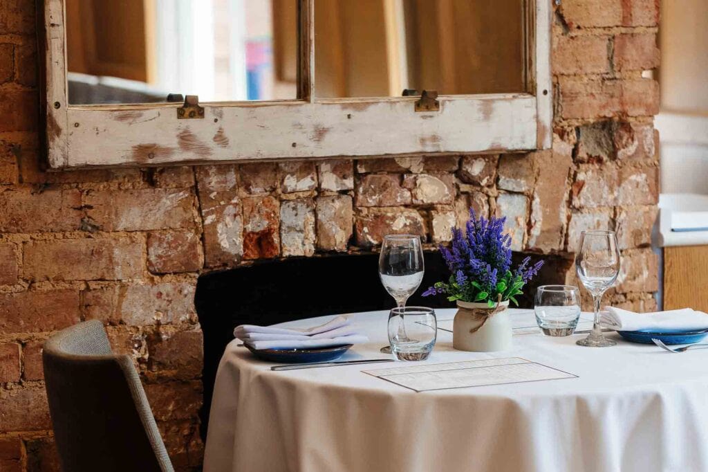 Elegant dining setup with a round table, white tablecloth, blue flowers, and glassware in a rustic brick-walled restaurant, perfect for food photography in Rutland.