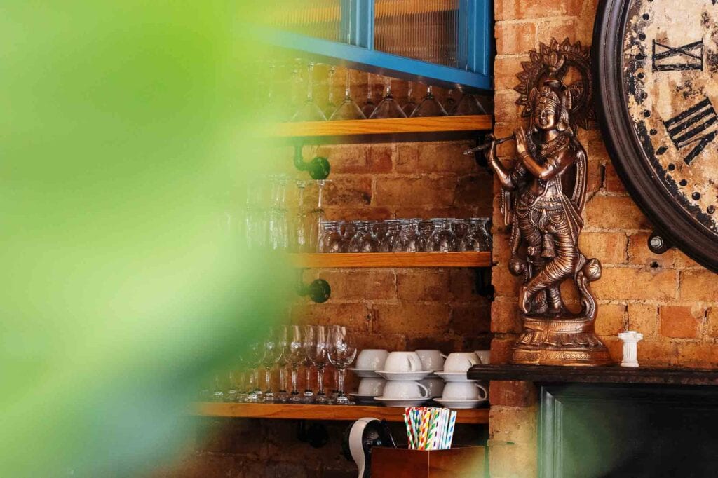 Interior shot of a rustic bar featuring a vintage clock, wooden shelves with glassware, and a decorative statue, showcasing warm brick walls and eclectic pub decor.