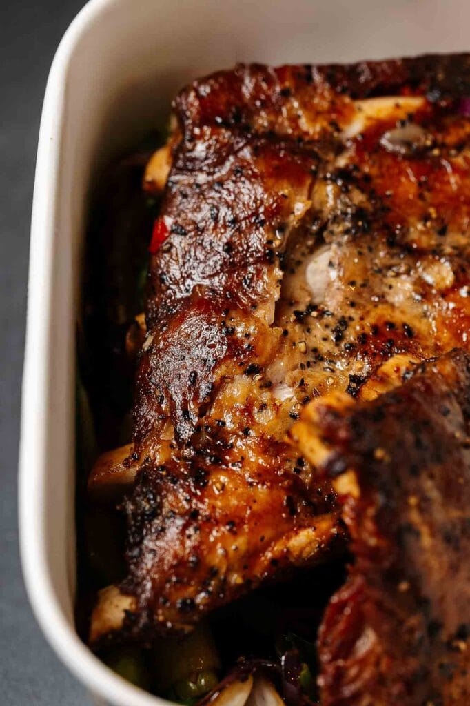 Close-up of a perfectly roasted lamb shoulder with crispy skin, seasoned with herbs and spices, served with roasted vegetables in a white ceramic dish.