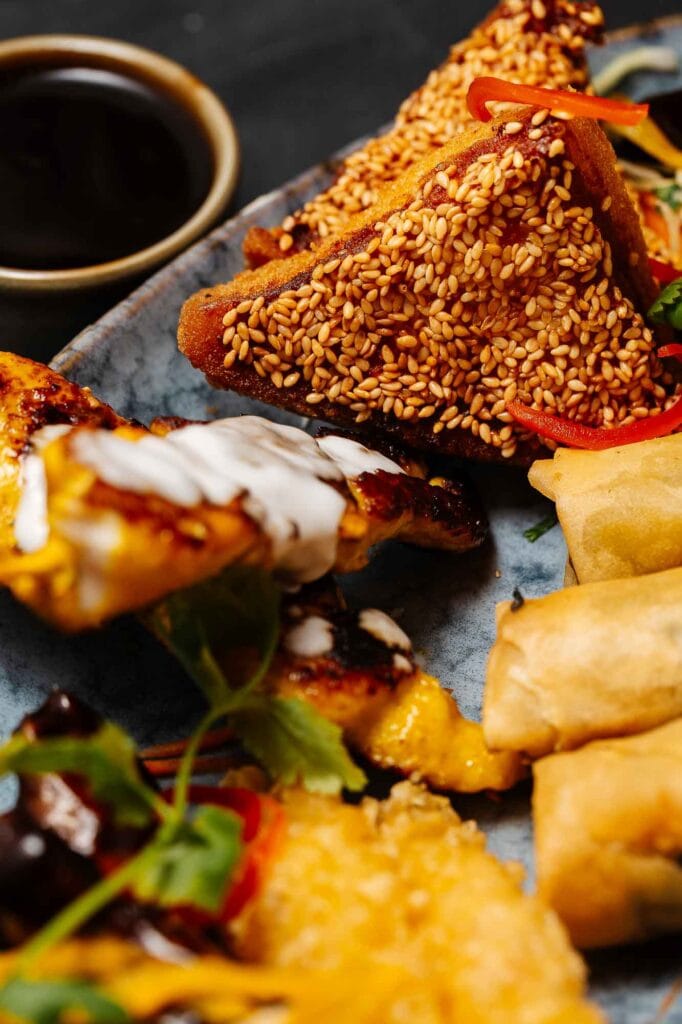 Brightly coloured sesame chicken bites with crispy coating and fresh vegetable garnish, served with dipping sauce on a rustic plate.
