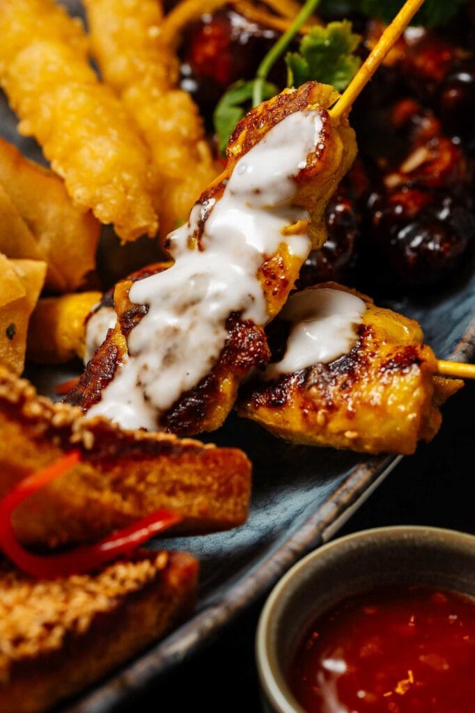 Delicious grilled chicken skewers topped with creamy sauce, served alongside crispy fried foods and fresh vegetables, perfect for food photography showcasing vibrant textures and flavours.