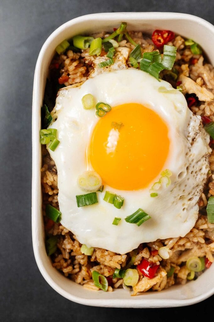 Delicious stir-fried rice topped with a perfectly cooked sunny-side-up egg, garnished with chopped spring onions and red peppers, served in a white bowl.