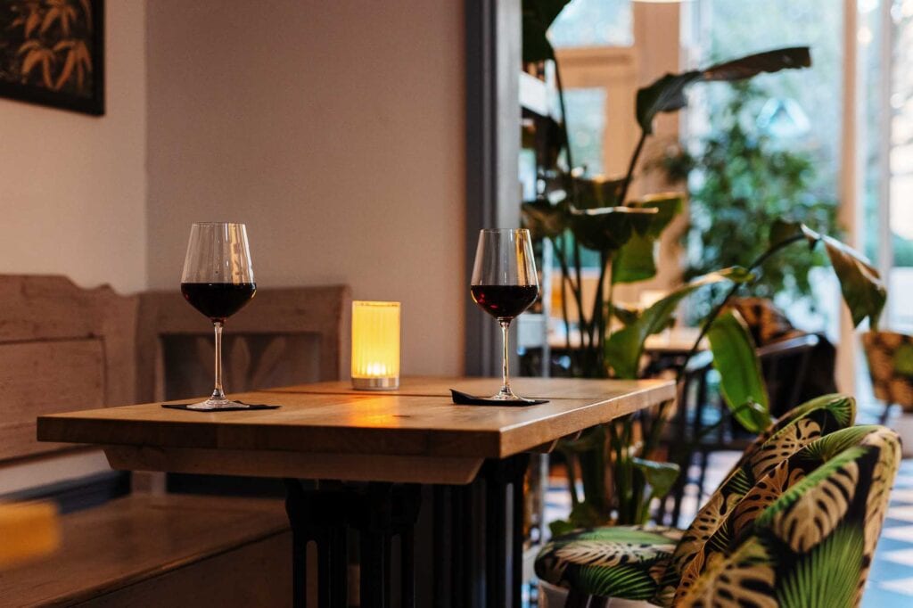 Two wine glasses filled with red wine on a wooden table in a warm, inviting restaurant with lush green plants and natural light.