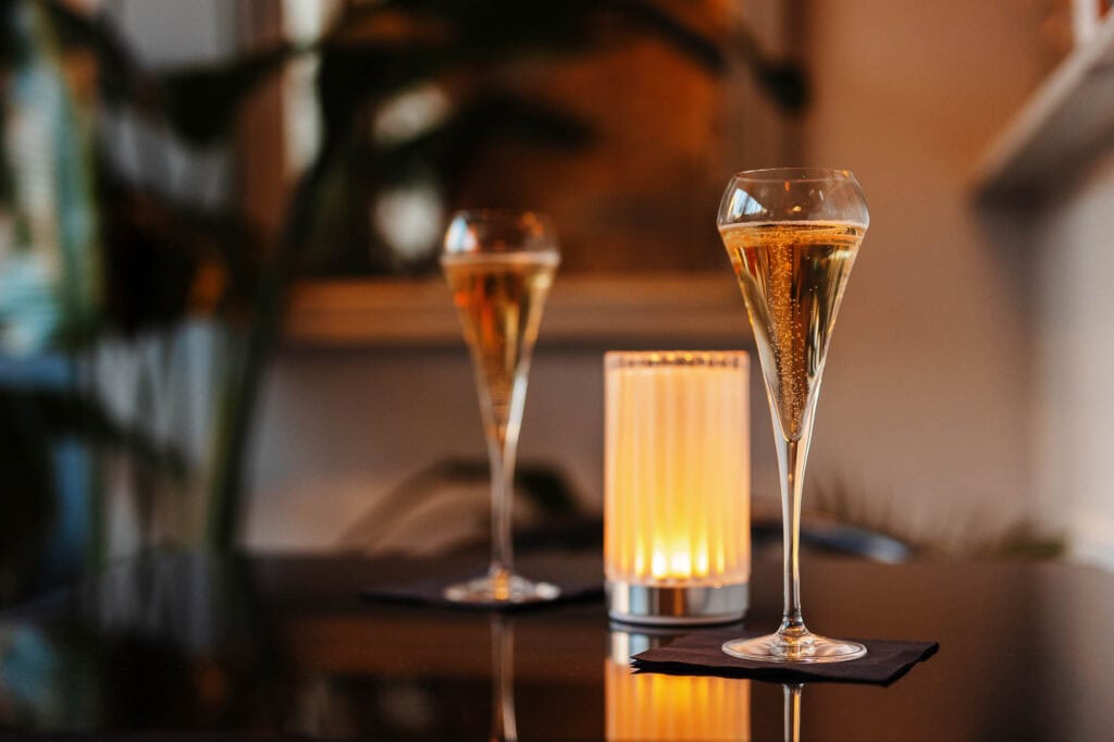 Two elegant champagne flutes filled with sparkling wine on a table, with warm ambient lighting in the background, showcasing professional food and beverage photography.