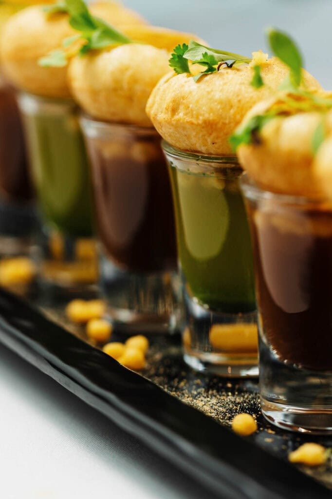 Delicious Indian-inspired dessert presentation featuring chickpea puffs garnished with fresh herbs, served in small glass cups with vibrant sauces, perfect for Indian restaurant menus and food photogr.