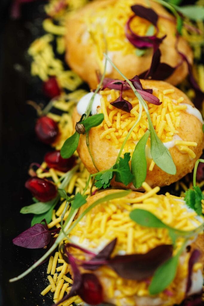 Close-up of Indian-inspired street food tacos topped with shredded cheese, fresh herbs, and microgreens, served on a black platter.
