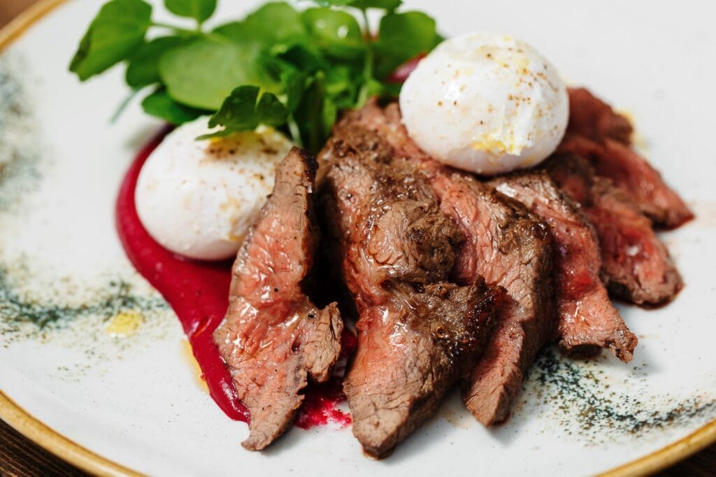 A close-up of perfectly cooked roast beef slices served with poached eggs, fresh greens, and a vibrant red sauce, showcasing high-quality food photography for culinary presentation.
