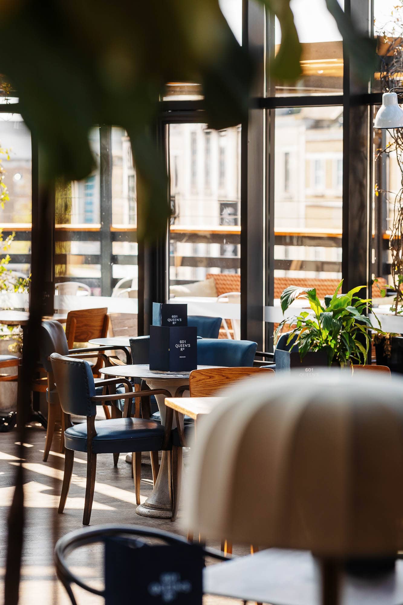 Bright and inviting café interior with large windows, wooden furniture, and lush green plants, perfect for relaxing or working in a stylish, contemporary setting.