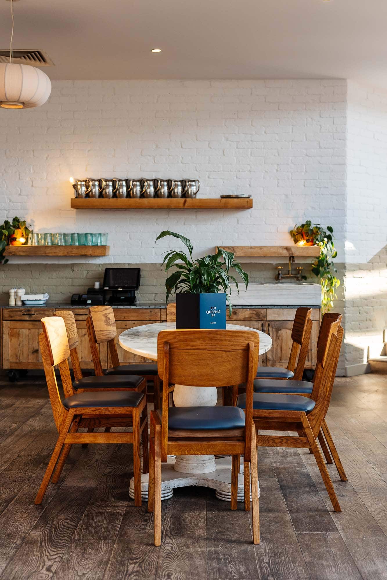 Modern rustic dining space featuring a round wooden table, eight matching chairs, and decorative shelves with plants and glassware in a bright, airy restaurant setting.