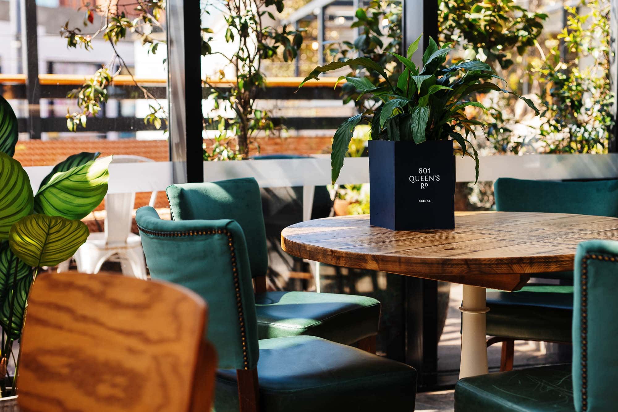 A stylish indoor dining space featuring a round wooden table surrounded by teal upholstered chairs, with lush plants and natural light creating a welcoming atmosphere.