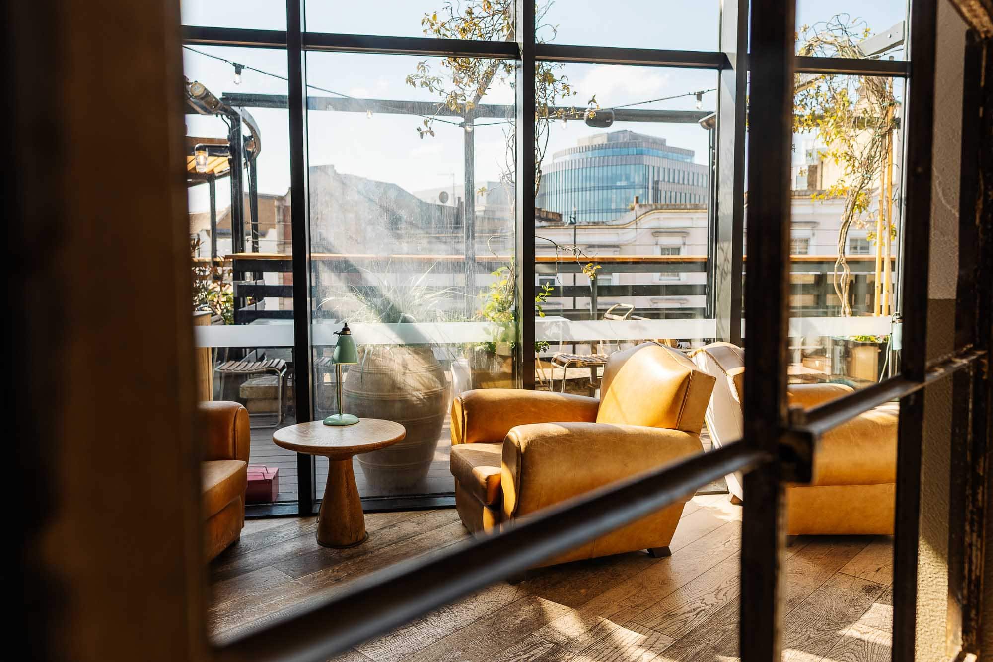 Bright rooftop lounge featuring comfortable mustard-yellow armchairs and a small round table, illuminated by natural sunlight streaming through large glass windows.