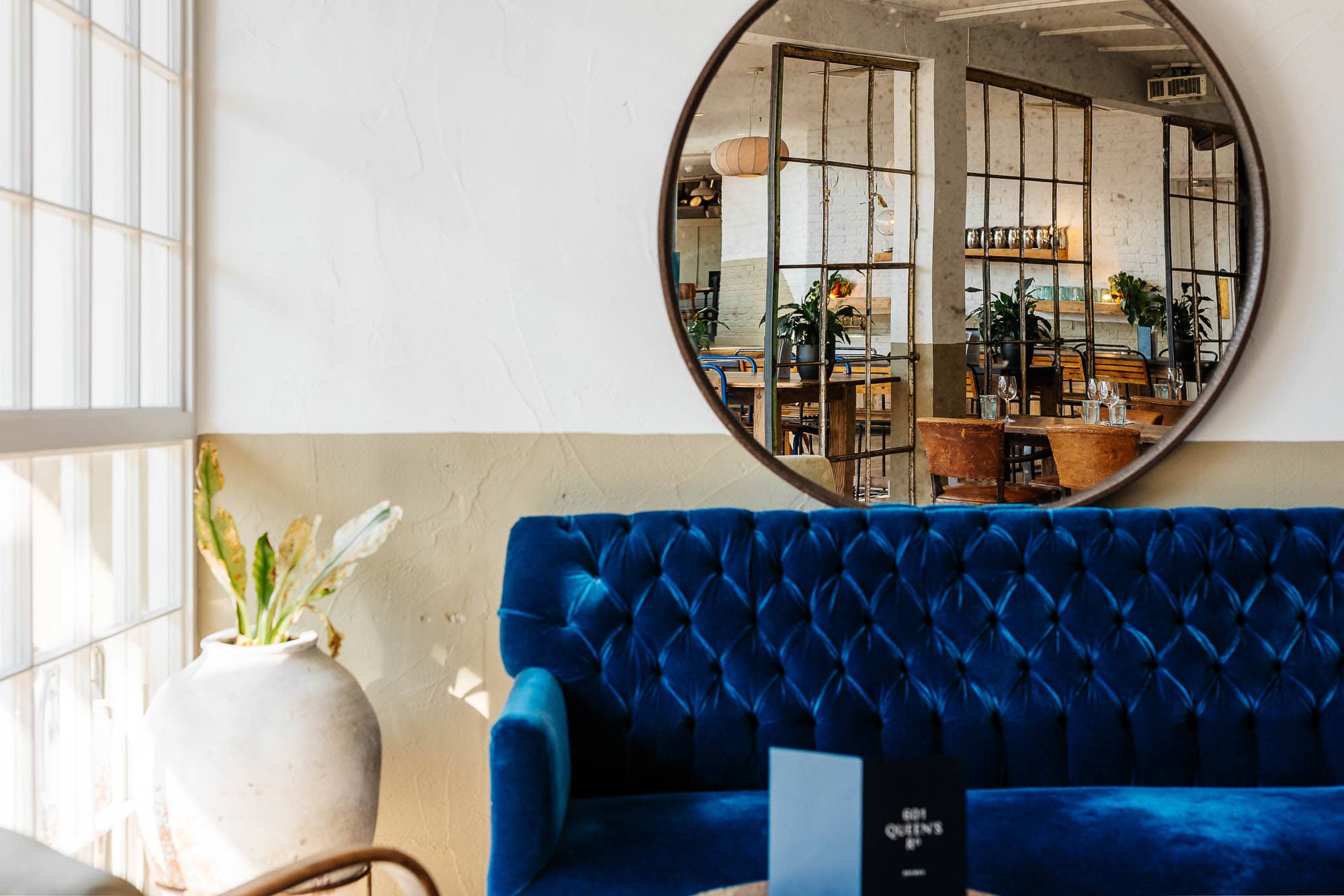 Elegant restaurant interior with contemporary design and stylish decor, featuring a plush blue velvet sofa, large round mirror, and natural light creating a welcoming atmosphere.